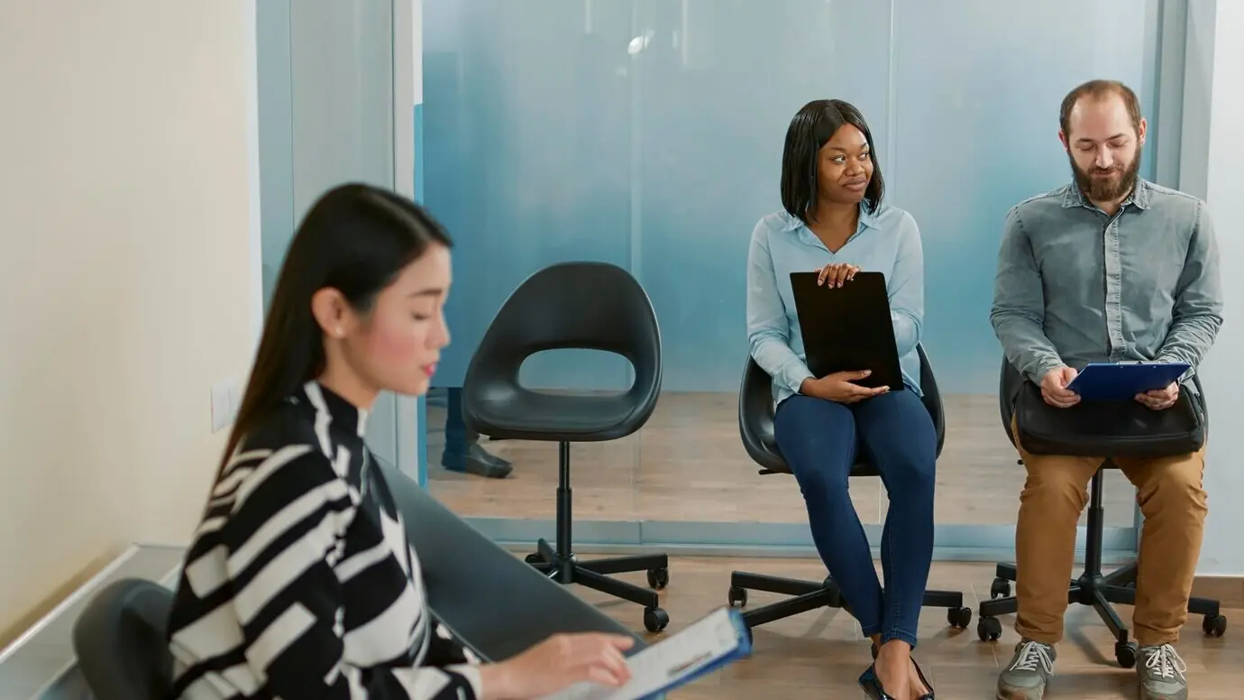 A diverse group of candidates converse in an office lobby, waiting to attend a job interview appointment. A man and a woman discuss the career opportunity and the application with a CV/resume.